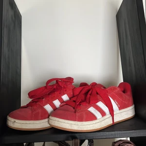 Röda Adidas Campus sneakers mocka - Snygga röda Adidas Campus sneakers i mocka med klassiska vita ränder och logga på hälen. Skorna har platt gummisula, rund tå och breda röda skosnören. Perfekta för dig som gillar retrostil och vill sticka ut med färg.