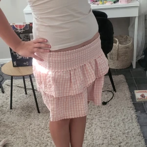 Rosa rutig volangkjol med smock - Supersöt kort kjol i ljusrosa och vitt med rutigt mönster. Kjolen har bred smock i midjan och dubbla volanger som ger en flowy känsla. Perfekt för varma dagar och lätt att matcha med en enkel topp. Tillverkad i mjuk bomull som känns skön mot huden. På första bilden är kjolen nervikt💕