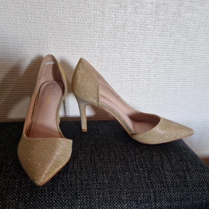 Glittriga guldiga pumps från Tiamo - Snygga pumps i guld med glittrig finish från Tiamo. Skorna har spetsig tå, smal klack och ett elegant randigt mönster i materialet. Perfekta för att sticka ut och ge din outfit extra bling. Klassiskt snitt med öppen insida och bekväm passform. Nypris 799, använda en gång. 
