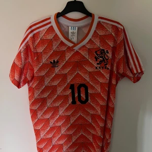 Holländsk retro fotbollströja Adidas - Säljer en ikonisk orange fotbollströja från Adidas med KNVB-logga och nummer 10. Tröjan har korta ärmar, vit-orange V-ringning och klassiska vita ränder på axlarna. Mönstret är geometriskt och riktigt retro, perfekt för dig som älskar klassisk fotbollsstil.