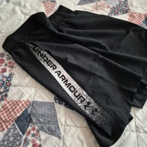Under Armour shorts - Svarta träningsshorts från Under Armour med vit logga och tryck längs ena benet. Modellen är loose fit med elastisk midja och snörning på insidan. Perfekta för gymmet eller löprundan, tillverkade i lätt och snabbtorkande syntetmaterial.