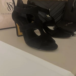 Svarta klackade sandaler i mocka - Stilrena svarta sandaler med hög klack och öppen tå från SO All. Skorna är gjorda i mjuk mocka med snygga utskurna detaljer och en stabil träfärgad klack. Perfekta för dig som vill ha en edgy men elegant look.