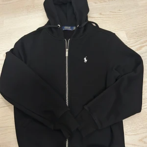 Svart Ralph lauren zip - Svart hoodie från Polo Ralph Lauren med dragkedja framtill och klassisk broderad logga på bröstet. Tröjan har huva med snörning och ribbade muddar vid ärmslut och nederkant. Perfekt för en chill och stilren look.