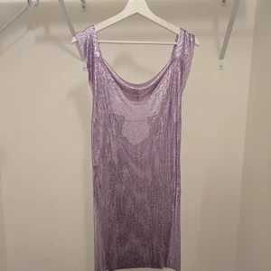 Lila glittrig kortklänning i metall - Superfin lila glittrig kortklänning i metall med ärmlös design och snygg draperad urringning. Klänningen har ett mesh-liknande material som ger en cool och skimrande effekt. Perfekt för dig som vill sticka ut och glänsa.