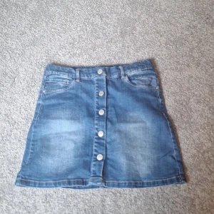 Blå jeanskjol med knappar från Name It - Snygg blå jeanskjol från Name It med knappar hela vägen framtill. Kjolen har klassiska fickor både fram och bak samt hällor för bälte. Endast använd en gång! Pris kan diskuteras.