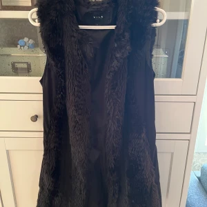 Svart pälsväst från Vila, - Säljer en svart pälsväst från Vila i storlek M. Västen har mjuk fuskpäls fram och bak och släta sidor. Går även att knyta i ryggen så att den passar bättre💕