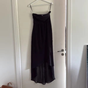 Svart långklänning från Vero Moda - Svart långklänning från Vero Moda med tunna axelband och snygg omlottliknande överdel. Klänningen har asymmetrisk nederkant med längre bakstycke och är tillverkad i ett lätt, flytande tyg som ger en elegant siluett.