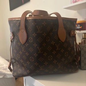  Monogram axelväska - Säljer en klassisk axelväska i brunt monogrammönster med ljusbruna skinnhandtag och detaljer. Väskan har rymlig insida, guldfärgade metalldetaljer och ikoniska loggor över hela ytan. Perfekt för dig som vill ha en tidlös och lyxig accessoar.