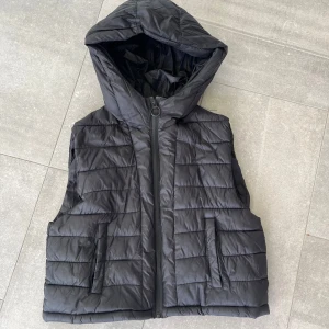 Svart dunväst med huva från Zara - Snygg svart dunväst från Zara med quiltad design och stor huva. Västen har dragkedja framtill och två fickor på sidorna. Perfekt för lager-på-lager och enkel att matcha med olika outfits.