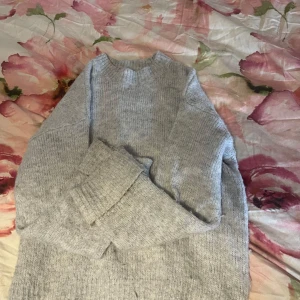 Ljusgrå stickad tröja med ribbade muddar - Mysig ljusgrå stickad tröja med rund hals och ribbade muddar vid ärmslut och nederkant. Tröjan har en enkel och clean look, perfekt för lager-på-lager och chill dagar. Materialet känns mjukt och värmande, utsvängda armar. Fläcket är sminkfläck går att ta bort.