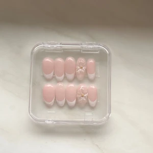 Press-on naglar med fransk design - Snygga press-on naglar i ljusrosa med vit fransk tipp och två naglar dekorerade med 3D-blommor och gulddetalj. Kommer i en genomskinlig ask och inkluderar nagellim. Perfekt för dig som vill ha en clean och söt look utan att gå till salong.