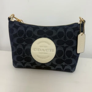 Blå axelväska från Coach med logomönster - Snygg axelväska från Coach i mörkblått tyg med ikoniskt logomönster och detaljer i beige skinn. Väskan har guldfärgade metalldetaljer, dragkedja upptill och en rund Coach-patch framtill. Perfekt storlek för det viktigaste och en stilren look.