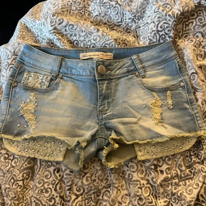 Ljusa jeansshorts med pärlor & spets - Snygga ljusblå jeansshorts från Monday denim med slitna detaljer, pärlor och spets på fickorna. Rå kant nertill och coola rivna partier framtill. Perfekta för sommaren och ger en trendig vibe till din outfit.