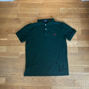 Mörkgrön piké från Polo Ralph Lauren - Mörkgrön slim fit pikétröja från Polo Ralph Lauren med klassisk krage, tre knappar och röd broderad logga på bröstet. Tillverkad i mjuk bomull och har korta ärmar. Perfekt för en clean och stilren look.