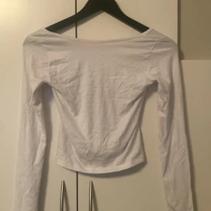 Vit långärmad topp från Gina Tricot - Enkel och stilren vit långärmad topp från Gina Tricot. Modellen har en bred, rundad halsringning och är figurnära i passformen. Tillverkad i mjuk bomull som känns skön mot huden. Perfekt att styla med jeans eller kjol för en clean look.