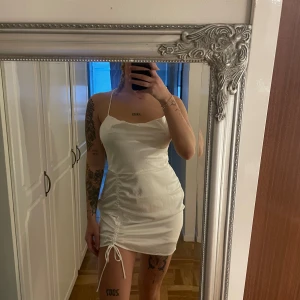 Vit kort klänning med snörning - Säljer en vit kort klänning med smala axelband och snygg snörning i sidan. Klänningen har öppen rygg med korsade band och en tight passform. Klänningen är i strl 40 men passar 38 då den går att anpassa med snörningen i ryggen. Använd en gång.