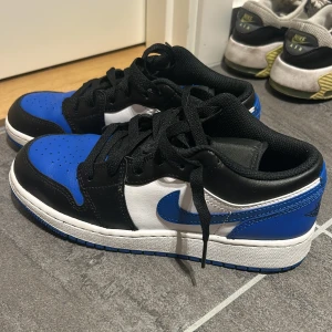 Nike Air Jordan 1 Low blå/svart/vit - Snygga Nike Air Jordan 1 Low sneakers i blått, svart och vitt skinn. Klassisk siluett med svart snörning, blå yttersula och ikoniska Jordan Wings-loggan på hälen. Perfekta för dig som vill sticka ut med en tidlös streetstyle. Endast använda 3 gånger. Värde 1100 kr.