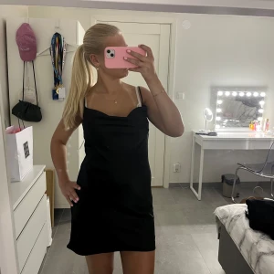 Svart satin klänning - Supersnygg svart kort klänning i satin med tunna axelband och lätt draperad urringning. Klänningen har en enkel och stilren design som ger en chic vibe. Perfekt att styla med smycken och klackar för en cool look.