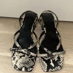 Svarta sandaler med ormmönster och klack - Trendiga sandaler med svart och vitt ormmönster, fyrkantig tå och smala remmar. Skorna har en stabil, låg klack och öppen tå, perfekt för att lyfta din outfit. Snyggt och edgy val för dig som gillar att sticka ut.