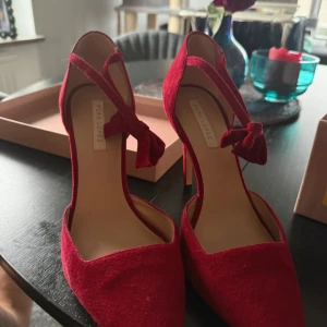 Röda mockapumps från Pura Lopez - Snygga röda pumps i mocka från Pura Lopez med spetsig tå och hög klack. Skorna har en elegant knytning runt vristen och är tillverkade i mjukt skinn invändigt. Perfekta för dig som vill sticka ut med en djärv färg och stilren design.