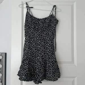 Svart blommig kortklänning  - Supersöt svart kortklänning från new yorker i storlek XS. Klänningen har vita små blommor, smala axelband som man kan justera själv och knyta och en lätt utställd kjol och den finns även shorts i den. Dragkedja i ryggen som funkar jättebra. Klännnngen är nästan inte använd. 