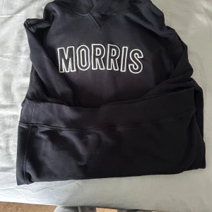 Svart hoodie från Morris XL - Svart hoodie från Morris i storlek XL med vit broderad logga på bröstet. Tröjan har huva med vita snören och en klassisk känguruficka framtill. Tillverkad i mjuk bomull, perfekt för en chill och stilren look.