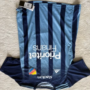 Djurgården fotbollströja Adidas M - Djurgården fotbollströja från Adidas i storlek M. Tröjan har klassiska blå och marinblå vertikala ränder, klubbmärke på bröstet och nummer 26 på ryggen. Tillverkad i lätt och ventilerande polyester, perfekt för match eller träning.