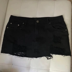 Svart lågmidjade jeanskjol   - Svart kort lågmidjade jeanskjol från NLY Jeans med slitna detaljer och fransig nederkant. Klassisk femficksmodell med knapp och dragkedja framtill. 