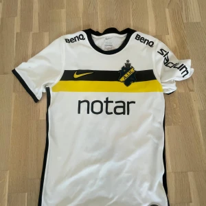 Vit AIK matchtröja Nike Milošević #5 - AIK:s officiella matchtröja i vitt med svarta och gula detaljer, tillverkad av Nike. Tröjan har korta ärmar, klubbmärke på bröstet, sponsortryck och Milošević samt nummer 5 på ryggen. Materialet är lätt och andas, perfekt för fotbollsträning eller match. ÄKTA priset är prutbart