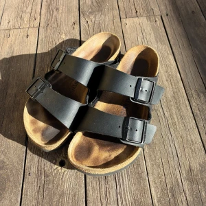 Birkenstock svarta - Klassiska svarta Birkenstock med två breda remmar i skinn och justerbara spännen. Fotbädden är formad för extra komfort och sulan har ett vågigt mönster för bra grepp. Perfekta för varma dagar och chill vibes.