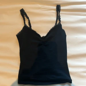 Svart spetslinnetopp från Bershka XS - Svart linne från Bershka i storlek XS med tunna axelband och v-ringning. Toppen har fina spetsdetaljer längs kanterna och en lätt rynkning vid bysten. Perfekt att styla med jeans eller kjol för en trendig look. Perfekt nu till sommaren🙌🏻💓