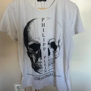  Philipp Plein t-shirt  - Vit t-shirt från Philipp Plein med stor svart dödskalle- och textprint på framsidan. Klassisk rund halsringning och korta ärmar. Tillverkad i mjuk bomull för en skön känsla. Perfekt för dig som gillar edgy streetstyle.