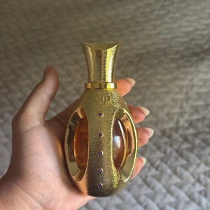 Oud Mood Gold parfymflaska - Lyxig parfym från Lattafa i en guldig, oval flaska med lila stenar och transparenta fönster som visar den bärnstensfärgade vätskan. Flaskan har en unik, texturerad yta och elegant form som verkligen sticker ut i parfymhyllan.