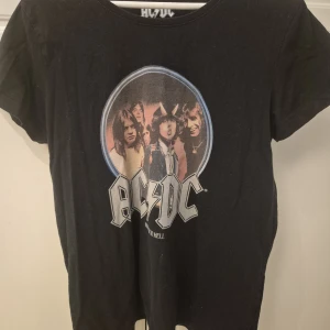Svart AC/DC t-shirt med tryck - Svart t-shirt med stort AC/DC-tryck och bandmedlemmar på framsidan. Klassisk rund hals och korta ärmar. Materialet känns mjukt och skönt, troligen bomull. Perfekt för dig som gillar rock och vill sticka ut med en ikonisk bandtröja.