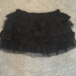 Svart volangkjol i tyll - Supersöt svart kortkjol med flera lager volanger i tyll och resår i midjan. Perfekt för dig som vill ha en lekfull och trendig look. Kjolen har en fluffig känsla och passar till många olika stilar.