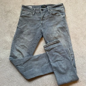 Grå jeans från Jack & Jones - Snygga grå jeans från Jack & Jones med klassisk femficksdesign och raka ben. Jeansen har en diskret tvättad look och är tillverkade i mjukt bomullsmaterial med en liten stretch för extra komfort. Perfekta för en avslappnad och stilren outfit. Modellen är Slim fit.
