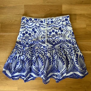 Vit och blå broderad kjol - Superfin kortkjol i vitt och blått med broderat mönster och dekorativa knappar framtill. Kjolen har hög midja och vågig nederkant som ger en unik look. Perfekt för dig som vill sticka ut med en färgstark och detaljerad stil. Kjolen är andvänd ett fåtal gånger och har inga tecken på andvändning, den ser som ny ut. Skriv till mig ifall du har några frågor!