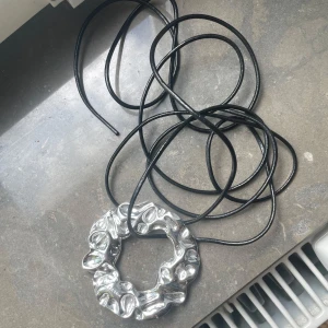 Silverfärgat halsband från Pilgrim - Unikt halsband från Pilgrim med en stor, rund och organisk formad silverfärgad hängare på svart läderrem. Hängaren har en cool, skulptural yta som verkligen sticker ut och ger edgy vibes. Perfekt för dig som gillar statement-smycken och vill ha något annorlunda.