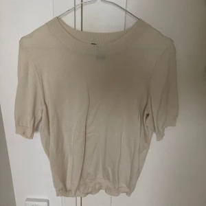Beige stickad kortärmad tröja Lindex - En stilren beige stickad tröja från Lindex i storlek S. Tröjan har korta ärmar, rund halsringning med bred ribbad kant och mjukt material som känns skönt mot huden. Perfekt för lager-på-lager eller som den är under vår och sommar.