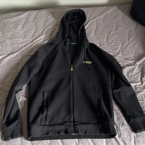 Svart hoodie från BOSS med dragkedja - Svart hoodie från BOSS i storlek M (passar S också) med guldfärgad dragkedja och logga på bröstet. Tröjan har huva med snörning, två fickor fram och ribbade muddar. 