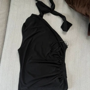 Svart one shoulder topp Gina Tricot - Snygg svart topp från Gina Tricot i one shoulder-modell med knytband vid axeln. Toppen har rynkade detaljer på sidan och är tillverkad i ett mjukt, stretchigt material som sitter skönt på kroppen. Aldrig använd men har tyvärr tagit bort prislappen