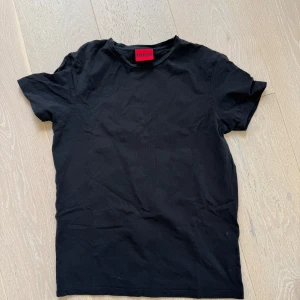 Svart t-shirt från Hugo, storlek S - En clean svart t-shirt från Hugo i klassisk modell med rund hals och korta ärmar. T-shirten är i mjuk bomull och har en diskret logga i nacken. Perfekt basplagg för en stilren look.