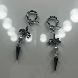 Silvriga örhängen med rosett och pärla - Unika silvriga örhängen med små rosetter, vita pärlor och coola konformade hängen. Ger en edgy men söt vibe och passar perfekt för dig som gillar att sticka ut. Säljs som par.