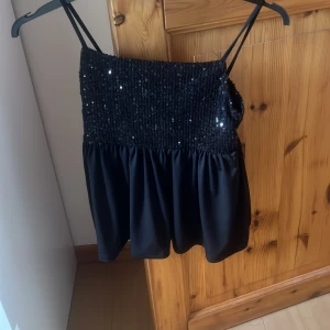 Svart paljett-tubtopp från Gina Tricot - Supersnygg svart tubtopp från Gina Tricot med smala axelband och glittriga paljetter upptill. Toppen har en smockad överdel och ett flowy tyg nedtill som ger en cool och festlig look. Perfekt för dig som vill sticka ut med lite extra bling!