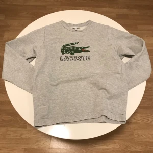 Grå tröja från Lacoste -  grå tröja från Lacoste med det ikoniska krokodilmärket på bröstet. Tröjan har långa ärmar och en rund halsringning, perfekt för en avslappnad look. Ett litet hål nere vid midje vecket