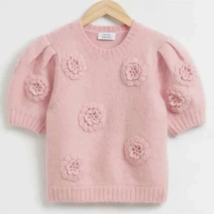 Rosa stickad blommig tröja & Other Stories - Supersöt ljusrosa stickad tröja från & Other Stories med korta puffärmar och virkade blommor som dekoration. Tröjan har rund halsringning och ribbade muddar. Perfekt för dig som vill ha en gullig och trendig look!