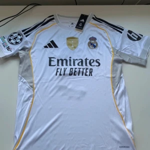 Real Madrid Mbappé matchtröja 2024 - Real Madrid 2024 matchtröja med Mbappé och nummer 9 på ryggen. Vit med svarta detaljer, guldiga inslag och gula linjer. Broderat klubbmärke, Adidas-logga och Champions League-märke på ärmen. Tillverkad i lätt funktionsmaterial (AEROREADY).