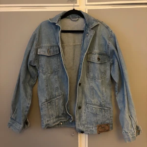 Ljusblå jeansjacka från Rolly, står ingen storlek men passar M - Snygg ljusblå jeansjacka från Rolly med klassisk passform. Jackan har fyra fickor framtill, knappar och coola sömdetaljer. Perfekt för dig som gillar en avslappnad och tidlös stil. Tuff patch med logga nertill på ena sidan.
