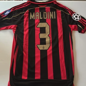 AC Milan Maldini #3 Champions League tröja - Säljer en klassisk AC Milan fotbollströja med Maldini och nummer 3 på ryggen. Tröjan är röd och svart med breda vertikala ränder, Champions League-patchar på ärmarna och guldfärgade detaljer. Tillverkad i lätt och ventilerande material, perfekt för dig som älskar fotbollshistoria. Replika. Endast handtvätt gäller!