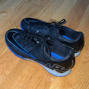 Nike Air Zoom svart/blå fotbollsskor - Snygga Nike Air Zoom fotbollsskor i svart med blå detaljer och blå yttersula. Skorna har snörning, låg profil och en sportig design med tydlig Nike-logga på sidan. Ovandelen är i syntetmaterial och sulan är mönstrad för bra grepp på planen.